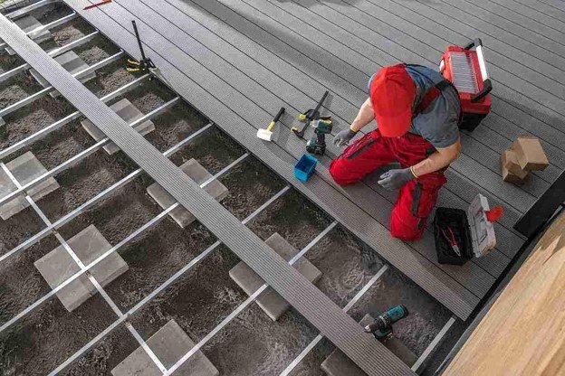 Worker in bright red overalls and red cap kneeling on grey composite deck installation.